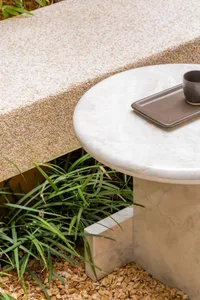 A round marble table with a tray and cup on a natural stone patio surrounded by greenery.