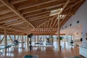 A spacious, modern interior of a health facility with wooden beams, plants, and fitness equipment.
