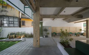 An industrial-style cafeteria with concrete flooring and walls, featuring green chairs and tables surrounded by plants.