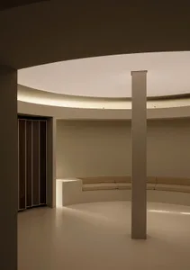 A minimalist interior space with a circular seating area and a central pillar, made of concrete.