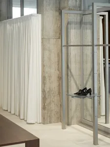 A modern fashion shop featuring a concrete wall, white curtains, and black shoes arranged on an aluminum shelf.