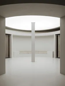 Modern minimalist room with white benches on a circular concrete platform, surrounded by white walls and a central white pill