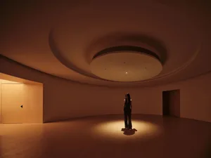 A minimalist interior with concrete walls and floors, featuring a single person standing in a spotlight.