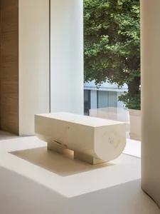 A minimalist wooden bench with a round section in front of a window with greenery outside.