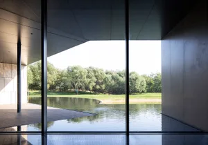 Modern building with glass walls overlooking a serene lake surrounded by greenery.