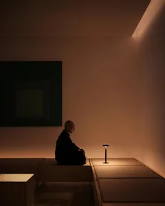 極簡室內，一人坐在昏暗房間，旁有燈具與一幅抽象畫框。