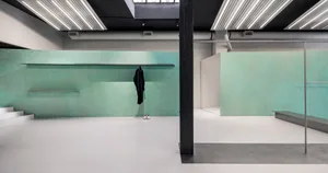 A minimalist gallery interior with a concrete floor, green textured walls, and a single black coat hanging from a shelf.