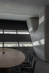 A modern conference room featuring a large round table surrounded by black chairs, with concrete walls and a large window pro