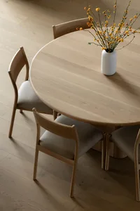 A round wooden dining table with chairs and a vase of flowers, set on a wooden floor, creating a minimalist interior design.