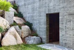 A modern entrance made of concrete with a large stone door and landscaping.