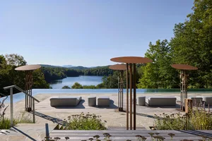 A modern outdoor living space featuring a wooden deck, infinity pool, and scenic lake view surrounded by lush greenery.