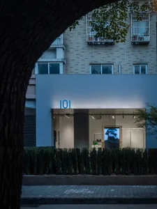 A modern retail storefront with a concrete exterior and green planters in front of a brick apartment building.