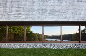 Modern residential building featuring a large glass window reflecting a serene lake surrounded by greenery, with a concrete f