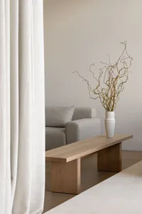 Minimalist living room featuring a wooden bench with a white vase of decorative branches and a gray sofa behind a white curta