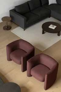 A modern living room with maroon fabric lounge chairs, a dark grey sectional sofa, and wooden tables on a light wood floor an