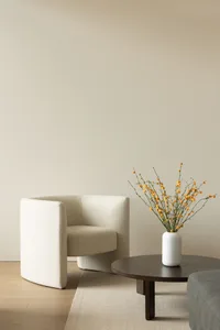 A minimalist living room with a white textured fabric armchair, round wooden coffee table, and a vase with yellow flowers.