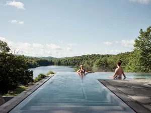 Two people relaxing in a modern outdoor infinity pool overlooking a serene lake and forested hills.