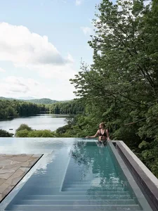 A woman in a swimsuit stands in an infinity pool overlooking a serene lake surrounded by trees and lush vegetation.