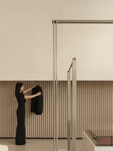 A woman in a minimalist living room, hanging a black coat on a metal drying rack. The room features concrete walls and floor,