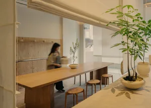 A modern restaurant interior features a long wooden table and stools, with plants and a minimalist design.