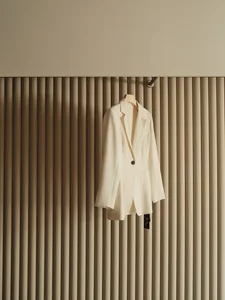 A white blazer hanging on a set of vertical beige curtains.