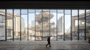 A photograph showing a large glass window in a construction site reflecting the surrounding urban landscape, including a mode