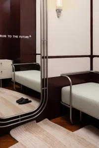 A minimalist hotel room featuring a metal frame bed with a light-colored mattress, placed on a wooden floor with a beige rug.