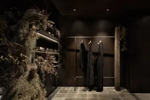 A modern clothing store with dark decor, featuring a wooden pillar, shelves with plants, and clothing on a hanging rail.
