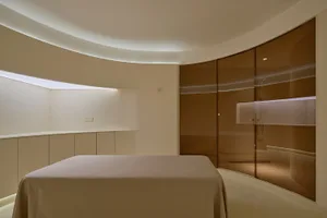 A minimalist bedroom with a glass wall and a large bed covered with a beige blanket.