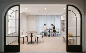 A modern waiting room with light-colored wooden flooring and minimalist decor, featuring two women seated at a table.