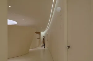 A modern office hallway with minimalistic design, white concrete walls, and a single person walking.
