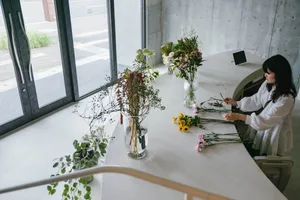 A woman sits at a white concrete counter arranging flowers using glass vases.