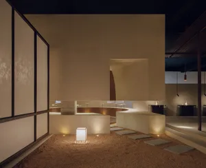 A minimalist gallery exhibition space featuring a sand-covered floor with sculptures and artistic lighting.