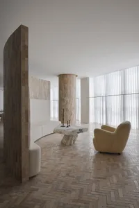 A modern living room featuring a large sofa, a stone table, and a fluffy armchair, set against a light-colored brick wall and