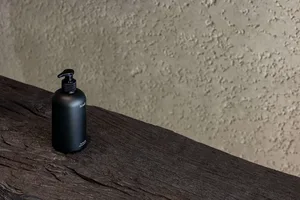 A black metallic bottle with a pump dispenser on a rustic wooden surface and a textured, beige background.