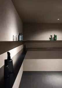 A minimalist bathroom with concrete walls and a black shelf holding various bottles and containers.