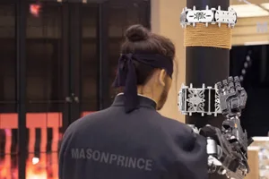 一位身穿背面印有「MASONPRINCE」字樣外套的人，正與安裝在立柱上的機械手臂互動。