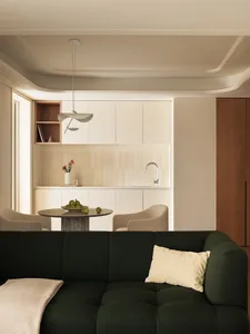 A modern living room featuring a green sofa, beige chairs, and a kitchenette with a sink and open shelving.