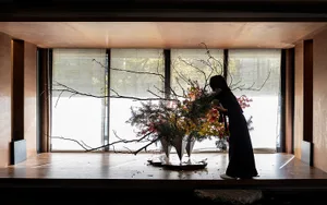 A woman in a black dress arranging a floral arrangement with wooden branches and foliage in a concrete space