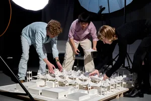 Three individuals working on a scale model of a building using paper and cardboard materials.