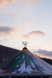 A colorful, triangular tent covered in snow with a weather vane on top.