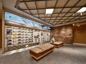 A retail shoe shop with a display of various shoes arranged on a wall, wooden benches, and a large screen showcasing shoe col