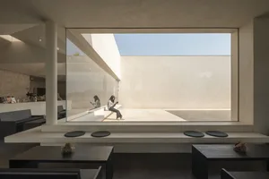 A minimalist coffee shop interior with a concrete wall and floor, featuring a large window and seating area.