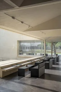 A spacious, modern cafe interior featuring concrete walls, high ceilings, and large windows overlooking a green courtyard.