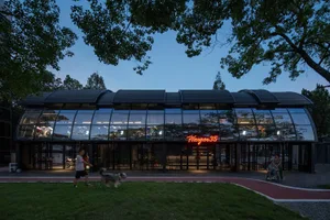 A modern restaurant exterior with large glass windows, illuminated signage, and a dog walking on the grass.
