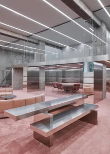 Modern office interior with minimalist design featuring metallic tables, glass partitions, and concrete pillars.