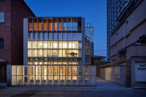 A modern glass facade building with illuminated interiors, set against a dusk sky.