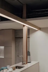 A minimalist kitchen with concrete walls and white countertops features a wooden beam with integrated lighting and a sleek st