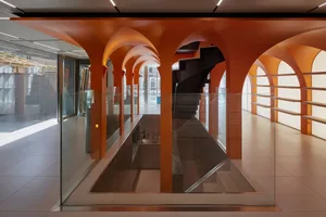 Modern interior space featuring a spiral staircase with metallic steps and orange columns under a glass railing.