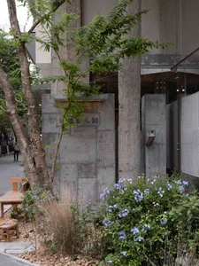 A modern building exterior with concrete columns, trees, and a flower bed with blue flowers.